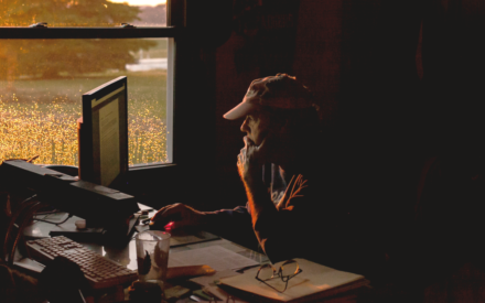 A farm reviews documents on his computer screen while sitting in a dark office. The sunset is seen through the window next to him.