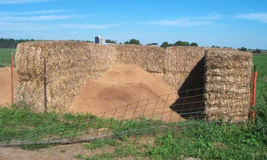 The image shows large stacks or walls of hay or straw bales in an agricultural field or farm setting. One of the stacks has an opening or passage cut through it, resembling an entrance or tunnel. The stacks are surrounded by wire fencing and green vegetation, indicating a rural, countryside location with an open sky in the background.